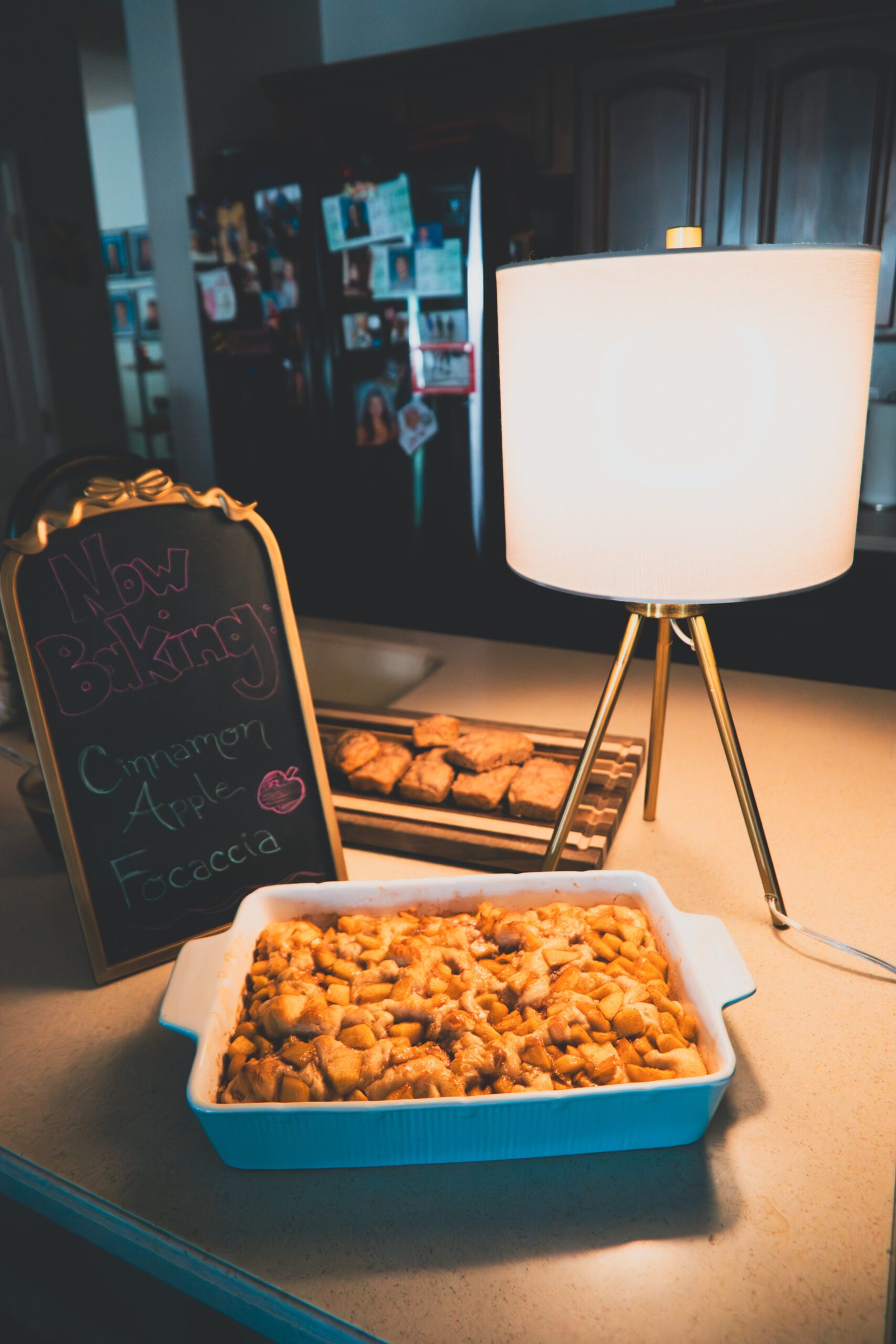 baker-01 Freshly baked cinnamon apple focaccia in a blue ceramic dish on a kitchen counter, next to a chalkboard sign reading 'Now Baking: Cinnamon Apple Focaccia' and a warm tripod lamp