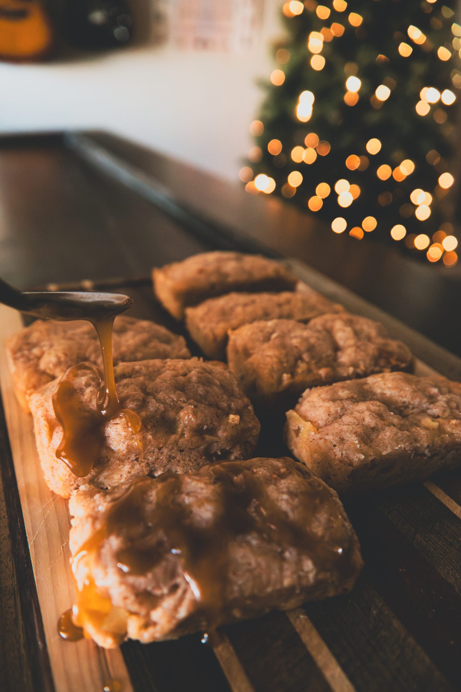 baker-03 Caramel sauce being drizzled from a spoon onto sliced cinnamon apple bread on a wooden cutting board, with Christmas tree lights glowing in the background
