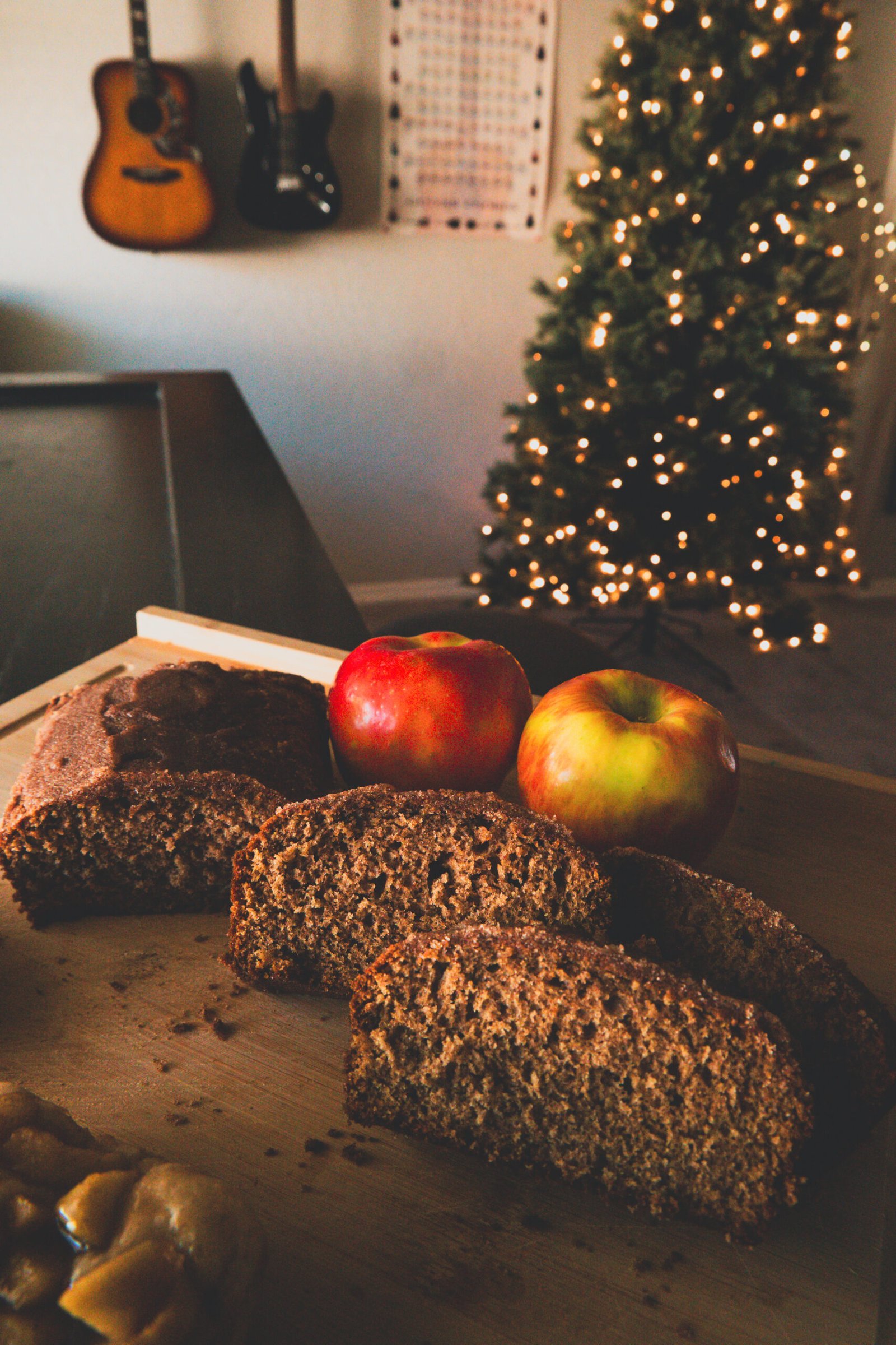 About Us 1 Caramel sauce being drizzled from a spoon onto sliced cinnamon apple bread on a wooden cutting board, with Christmas tree lights glowing in the background