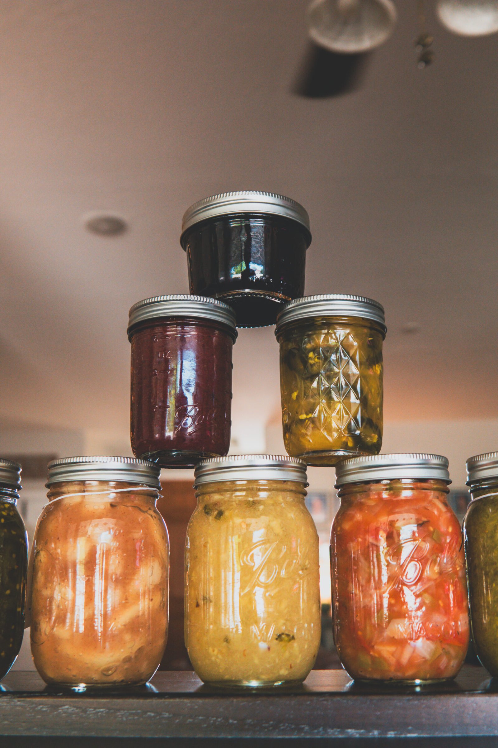 baker-12 Pyramid of colorful homemade canned goods in mason jars including berry jam, pickled jalapeños, relish, pickles, and stewed tomatoes